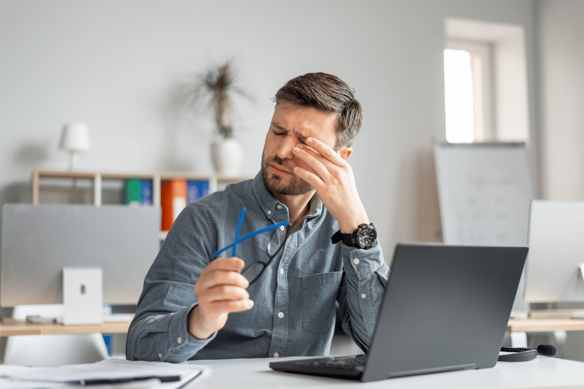 A man sitting in front of his computer and rubbing his eye due to discomfort
